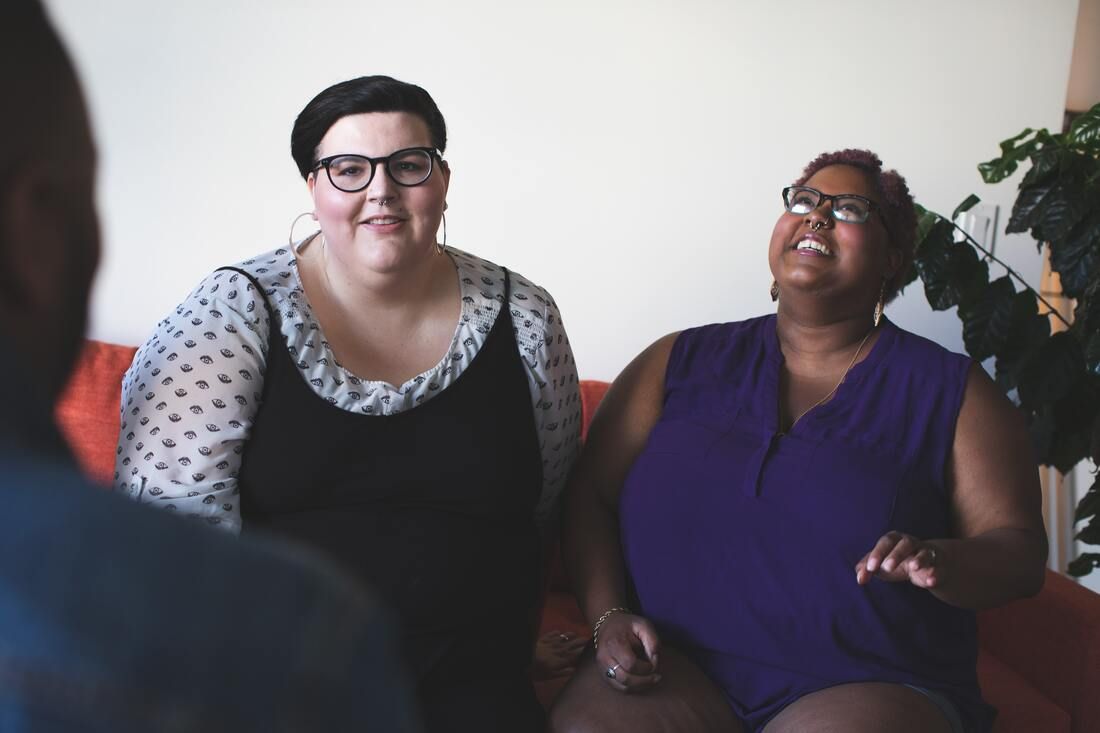 An interracial queer couple on a couch. The Black woman laughs, the white woman smiles.